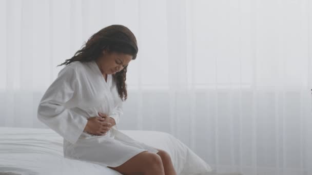 Female period pain. Young african american woman suffering from acute abdominal ache, sitting on bed, empty space — Stock Video