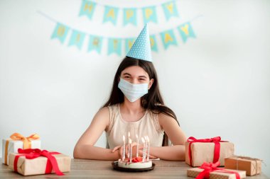 Parti şapkası ve yüz maskesi takan Hintli genç kız evde hediye kutuları ve doğum günü pastasıyla oturuyor.