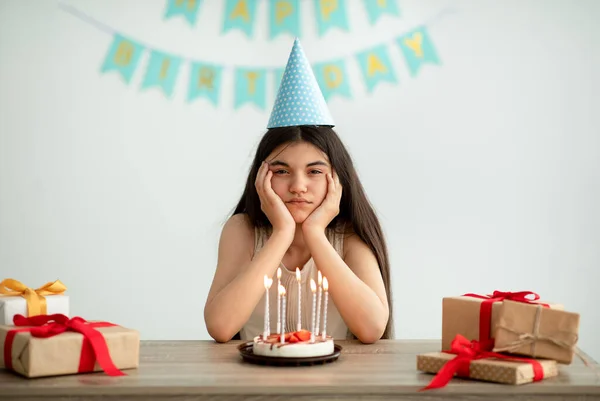 Hüsrana uğramış parti şapkalı genç kız portresi. Masada hediyeler ve doğum günü pastasıyla tek başına oturuyor. — Stok fotoğraf