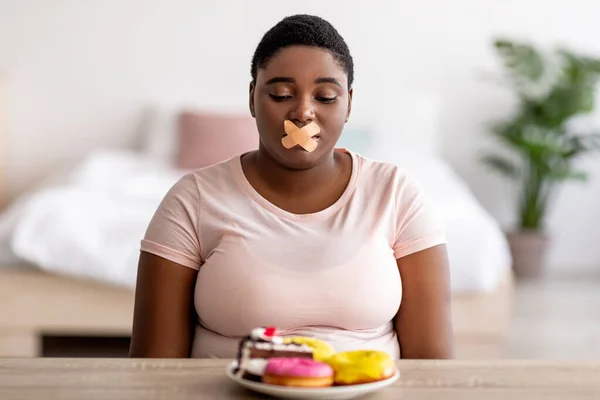 Potret wanita hitam gemuk yang tidak bahagia dengan perban perekat di mulutnya duduk di meja dengan sepiring manisan — Stok Foto