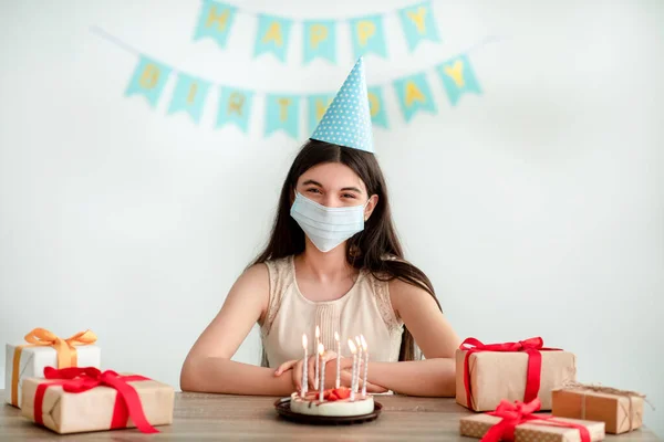 Parti şapkası ve yüz maskesi takan Hintli genç kız evde hediye kutuları ve doğum günü pastasıyla oturuyor. — Stok fotoğraf