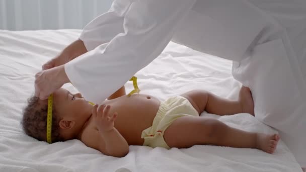 Medical nurse measuring head circumference of curly african american baby boy with tape, cute infant lying on bed — Vídeo de Stock