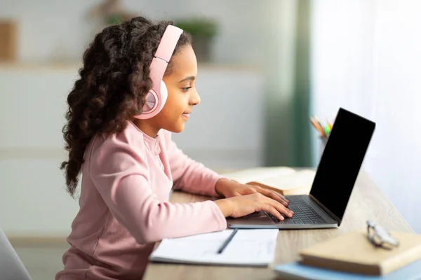 Bonito afro-americano menina adolescente ter on-line lição, usando laptop e fones de ouvido, sentado na mesa, vista lateral — Fotografia de Stock