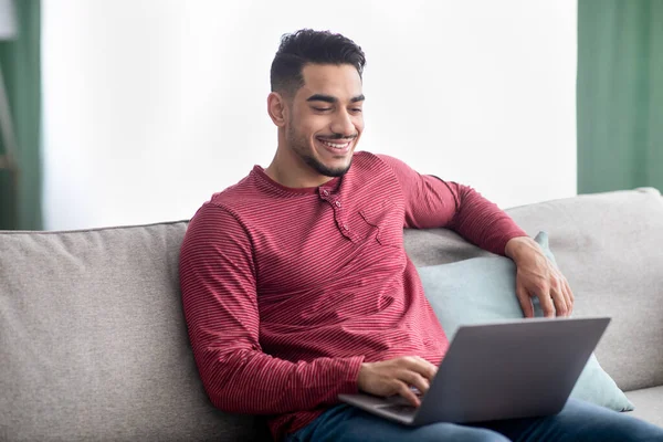 Fröhlich araber guy sitting auf couch at home, using laptop — Stockfoto