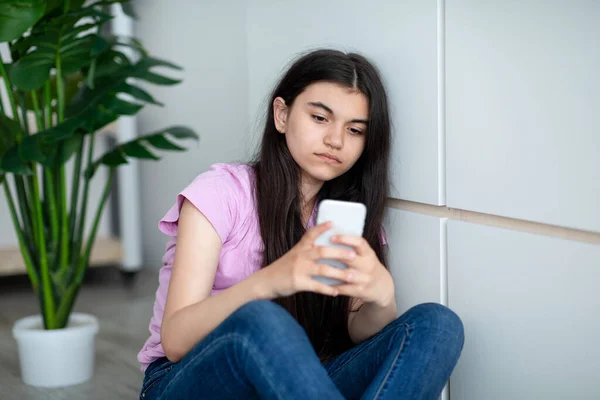 Depressieve Indiase tiener meisje met smartphone gevoel eenzaam, kijken naar het scherm, zitten in de buurt van de muur thuis — Stockfoto