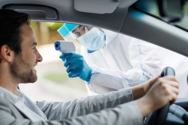 Pessoal médico em terno hazmat tomar temperatura para a febre para o homem antes covid-19 teste na estação drive thru no hospital — Fotografia de Stock