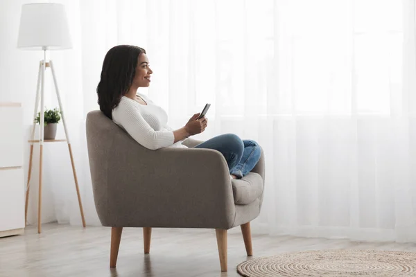 Relajada dama negra sentada en sillón y usando teléfono inteligente en casa, comprobando el correo electrónico, vista lateral, espacio para copiar — Foto de Stock