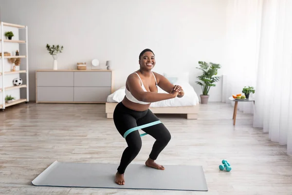 Longitud completa de la mujer negra con curvas en cuclillas con banda elástica durante el entrenamiento en casa, espacio de copia —  Fotos de Stock