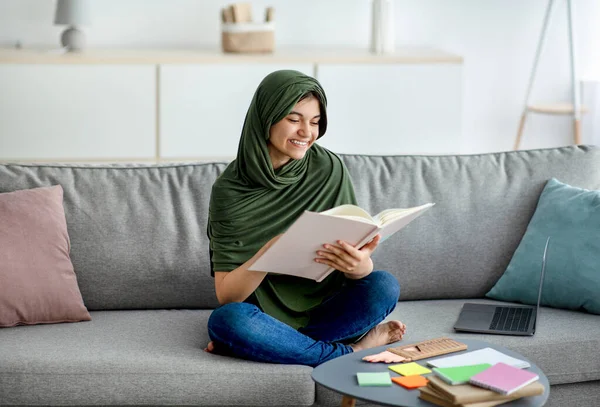 Happy teen girl in hijab sitting on sofa with textbook, reading, studying online from home, preparing for exam