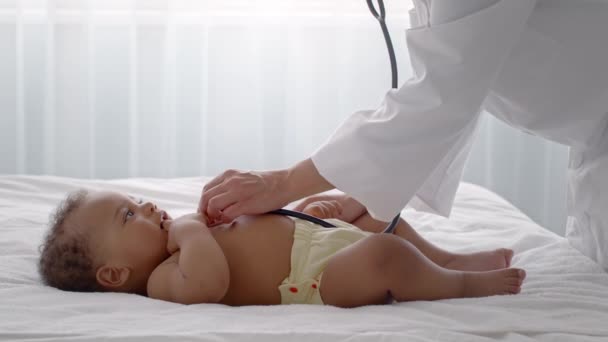 Lekarz pediatra badający małe dziecko stetoskopem w klinice, spokojne, kręcone afrykańskie niemowlę leżące na łóżku — Wideo stockowe