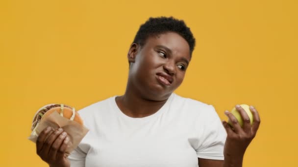 Fat Black Woman Choosing Burger Instead Of Apple, Yellow Background — Vídeo de Stock