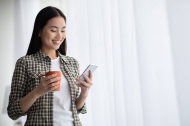 Elinde kahve fincanı olan güzel Asyalı kadın cep telefonu kullanıyor.