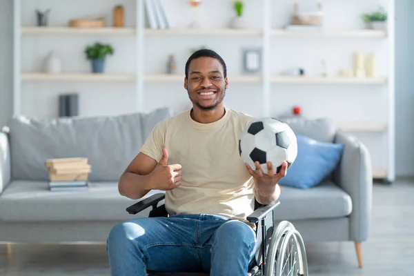 Vidám, fogyatékos, tolószékes fekete férfi, aki focilabdát tart, és hüvelykujját mutogatja otthon. — Stock Fotó