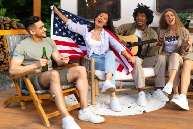 Gitar çalan, şarkı söyleyen, bira içen, elinde Amerikan bayrağı olan çok uluslu arkadaşlar kamp gezisinde karavanın yanında oturuyorlar.