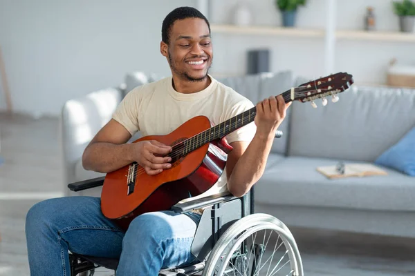 Boldog mozgássérült fekete srác tolószékben gitározik, zenét csinál a nappaliban. Maradj otthon tevékenységek során covid — Stock Fotó