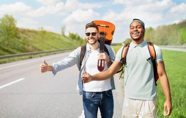 Dva chlápci s kytarovým stopováním na silnici, zastavení předjíždějících aut s palcem nahoru gesto, baví letní dobrodružství — Stock fotografie