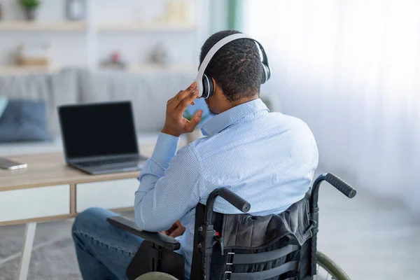 Visszapillantás a mozgássérült fekete srác tolószékben visel fejhallgató, használja laptop mockup design otthon — Stock Fotó