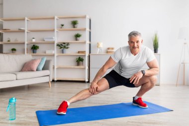 Kilitlenme ev antrenmanı. Oturma odasında yoga minderinin üzerinde bacaklarını esneten sportif olgun bir adam.