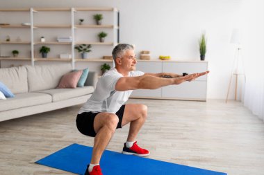 Çalışmak için evde tek başına kal. Spor giyimli, formda, oturma odasında çömelmiş, bacaklarını çalıştıran olgun bir adam.