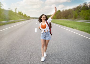Yaz macerası. Sırt çantalı neşeli genç bir kadın asfalt yolda yürüyor, heyecan içinde elini kaldırıyor.
