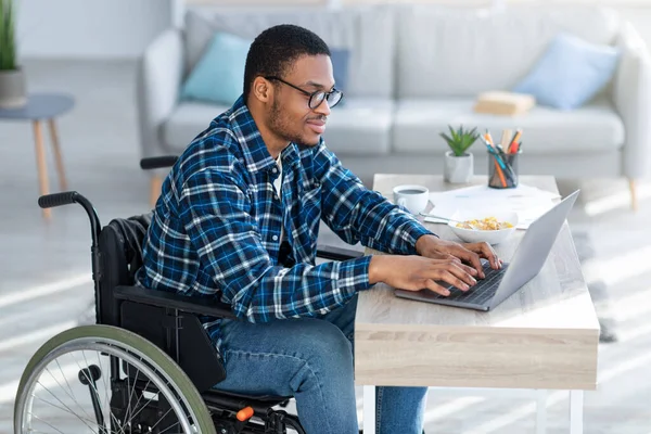 Online munka fogyatékkal élők számára. Mozgássérült fekete srác tolószékben, laptopot használ távoli otthoni munkára. — Stock Fotó