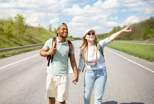 Cool joven pareja multiétnica cogidas de la mano mientras caminan por la carretera con mochilas, autoestop juntos —  Fotos de Stock
