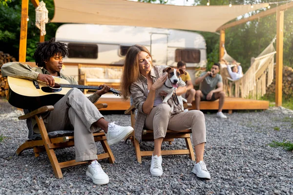 Young black guy playing guitar and singing, Caucasian lady holding dog near RV, camping with diverse friends in summer — Photo