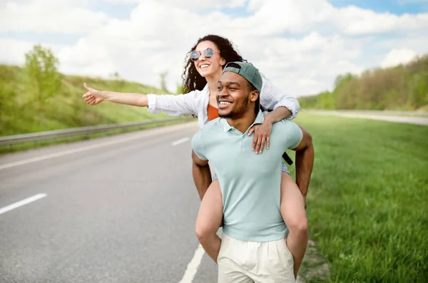 Chico negro fresco dando a su novia paseo a cuestas en la carretera, autoestop para el coche, mostrando el pulgar hacia arriba, detener el transporte —  Fotos de Stock