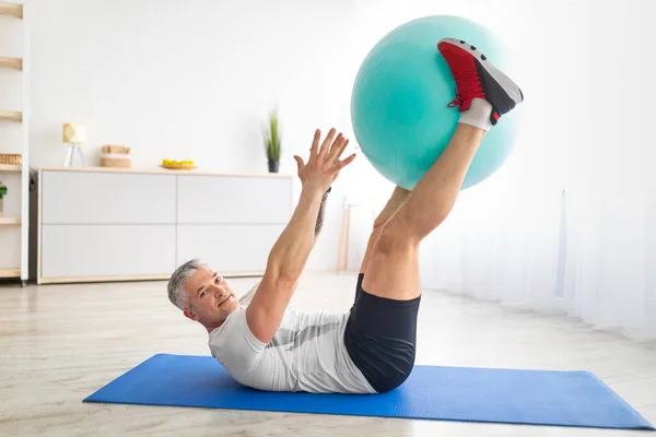 Esportivo homem maduro exercitando a imprensa muscular abdominal que coloca no chão com fitball atrás das pernas durante o treino matinal — Fotografia de Stock