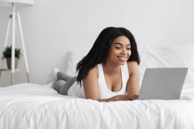 Yatak odasında iş, internette gezinme. Heyecanlı genç Afrikalı Amerikalı kadın dizüstü bilgisayarda çalışıyor, yatakta yatıyor.