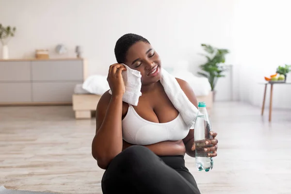 Plus Size Afroamerikanerin macht Pause vom häuslichen Training, hält eine Flasche Wasser in der Hand und wischt sich das Gesicht mit einem Handtuch ab — Stockfoto