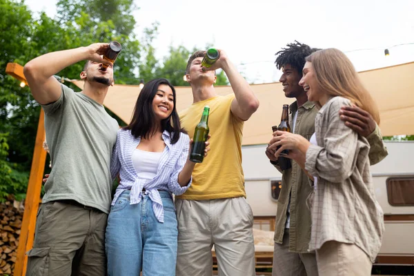 Cool multiracial young friends celebrating something, drinking beer, having social gathering or party near camper van