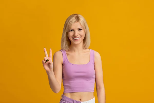 Buen humor. Mujer joven positiva mostrando victoria v signo, posando y sonriendo a la cámara, aislado sobre fondo amarillo —  Fotos de Stock