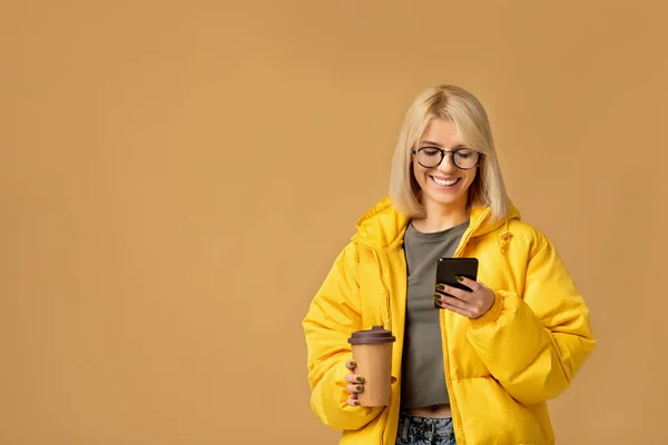 Τρόπος ζωής και κινητή τεχνολογία. Ευτυχισμένη κυρία με κίτρινο σακάκι γραπτών μηνυμάτων στο smartphone και πίνοντας καφέ takeaway — Φωτογραφία Αρχείου