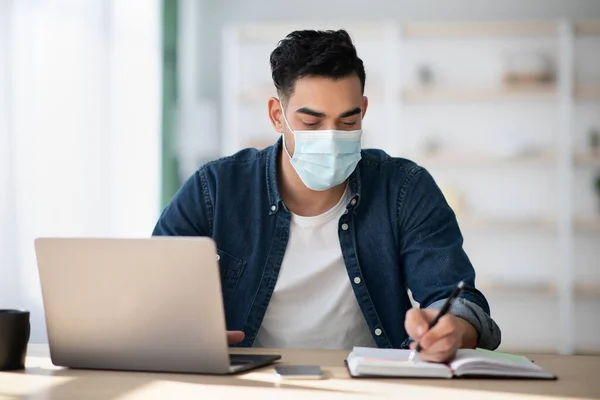 Homem árabe em máscara protetora usando laptop, tomando notas — Fotografia de Stock