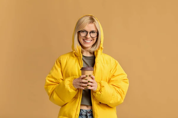 Гарячі напої. Позитивна молода леді в жовтій куртці тримає винос кави і посміхається камері, бежевий фон — стокове фото