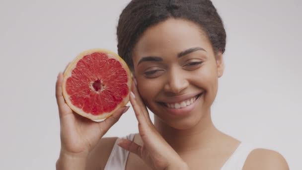 Vitaminas para cuidado de la piel y belleza. Mujer afroamericana bastante alegre sosteniendo media toronja cerca de su cara — Vídeo de stock