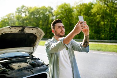 Kırık otomobilin yanında duran, cep telefonuyla el kaldıran, yol yardımını aramaya çalışan üzgün bir milenyum adamı.