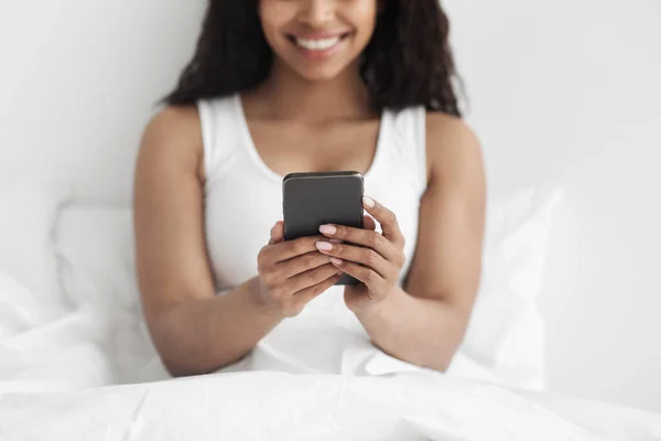 Feliz millennial africana americana dama relajante en cómoda cama, sosteniendo el teléfono inteligente en las manos, cosecha — Foto de Stock