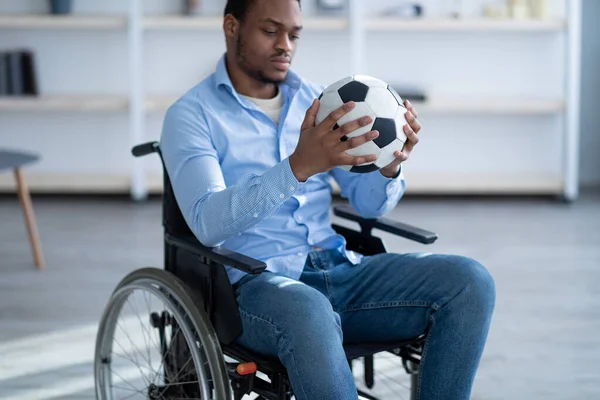 Mozgássérült fekete férfi focilabdával a kerekesszékben ülve, depressziósnak érzi magát a sérülése miatt otthon, szelektív fókusz — Stock Fotó