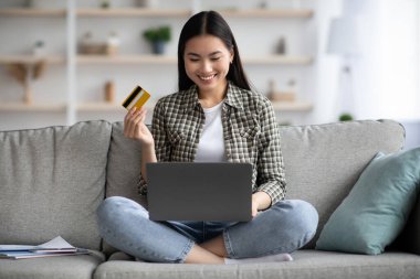 Evde dizüstü bilgisayar ve kredi kartı kullanan neşeli Asyalı kadın.