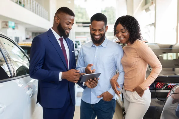 Vendedor de coches guapo que muestra los detalles de compra en la tableta digital a la pareja negra — Foto de Stock