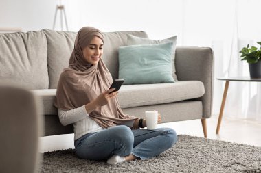 Bin yıllık mutlu Arap kadın tesettürlü kahve içiyor, sabah haberlerini akıllı telefondan okuyor.