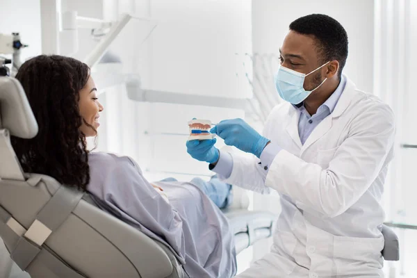 Svart stomatolog som holder kjeve av plast og underviser i tannhygiene for kvinnelige pasienter – stockfoto