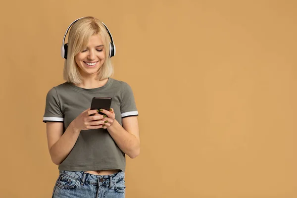 Calma senhora feliz vestindo fones de ouvido sem fio e usando smartphone, desfrutando de lista de reprodução de áudio favorito, fundo bege — Fotografia de Stock