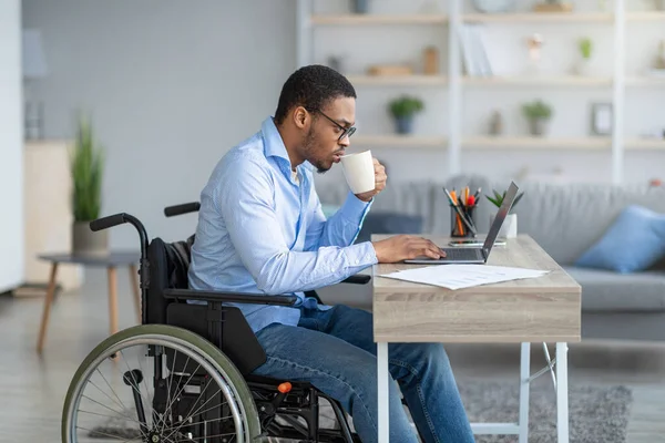 Súlyos fogyatékos fekete férfi tolószékben kávét iszik, videó leckét néz laptopon, online dolgozik otthonról — Stock Fotó