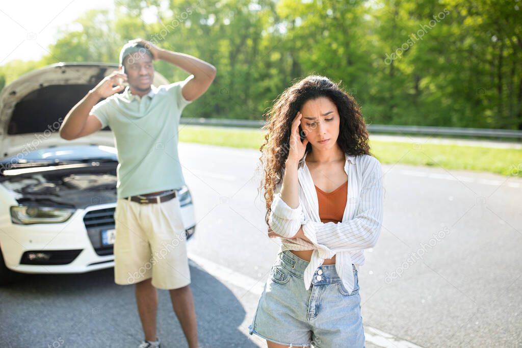 Young stressed woman standing on highway near broken car while boyfriend calling breakdown road service on smartphone