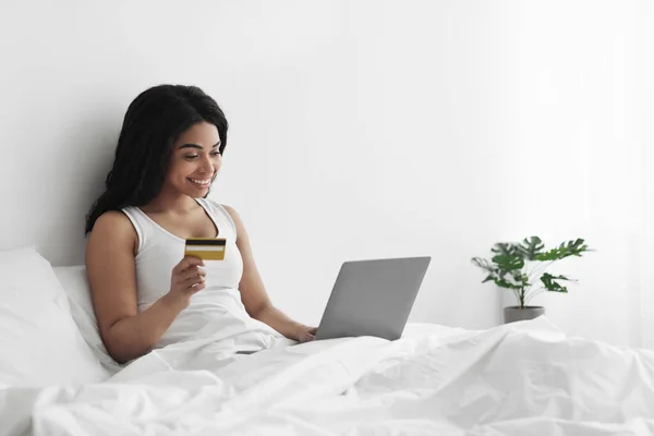 Concepto de comercio electrónico. Feliz joven afroamericano sentado en la cama, pagando con tarjeta de crédito a través de ordenador portátil, espacio libre — Foto de Stock
