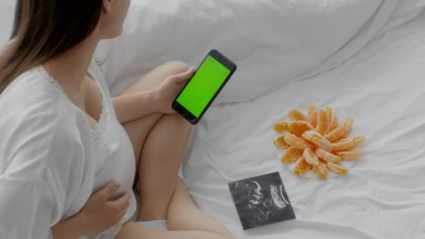 Una donna incinta è sdraiata a letto con un telefono in mano che le tiene lo stomaco, coprendo le gambe con una coperta, vista dall'alto — Video Stock