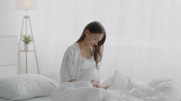 Bella mujer embarazada feliz sentada en la cama y abrazando el vientre — Vídeos de Stock